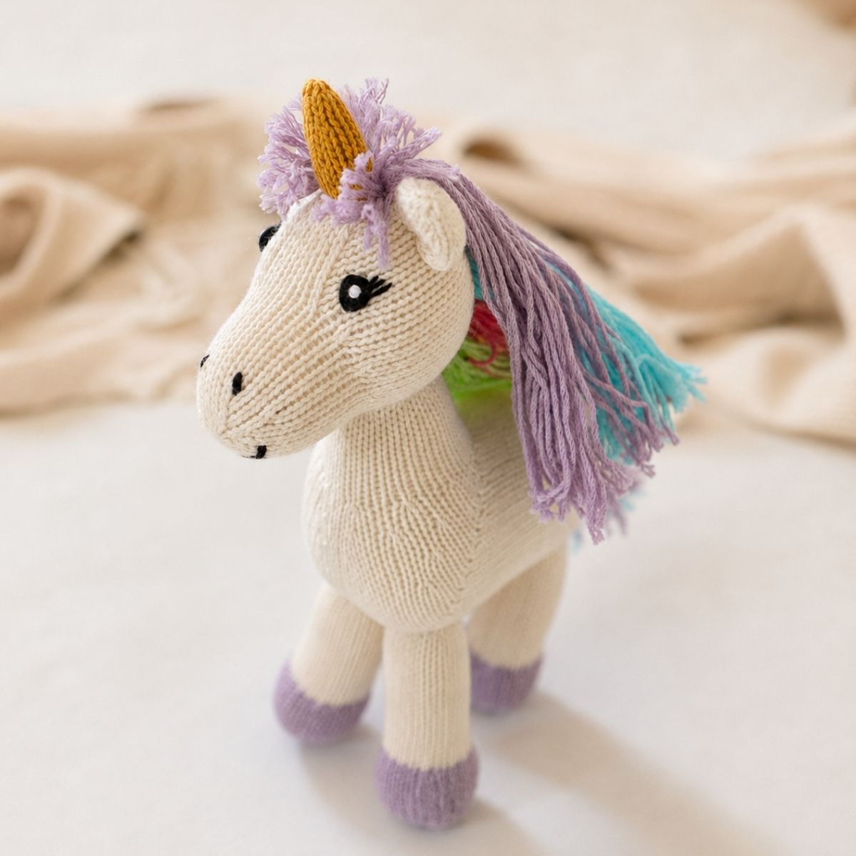 Vollständig abgebildetes hand handgemachtes Bio Kuscheltier Einhorn mit bunter Mähne, gestricktem Horn und lilafarbenen Hufen