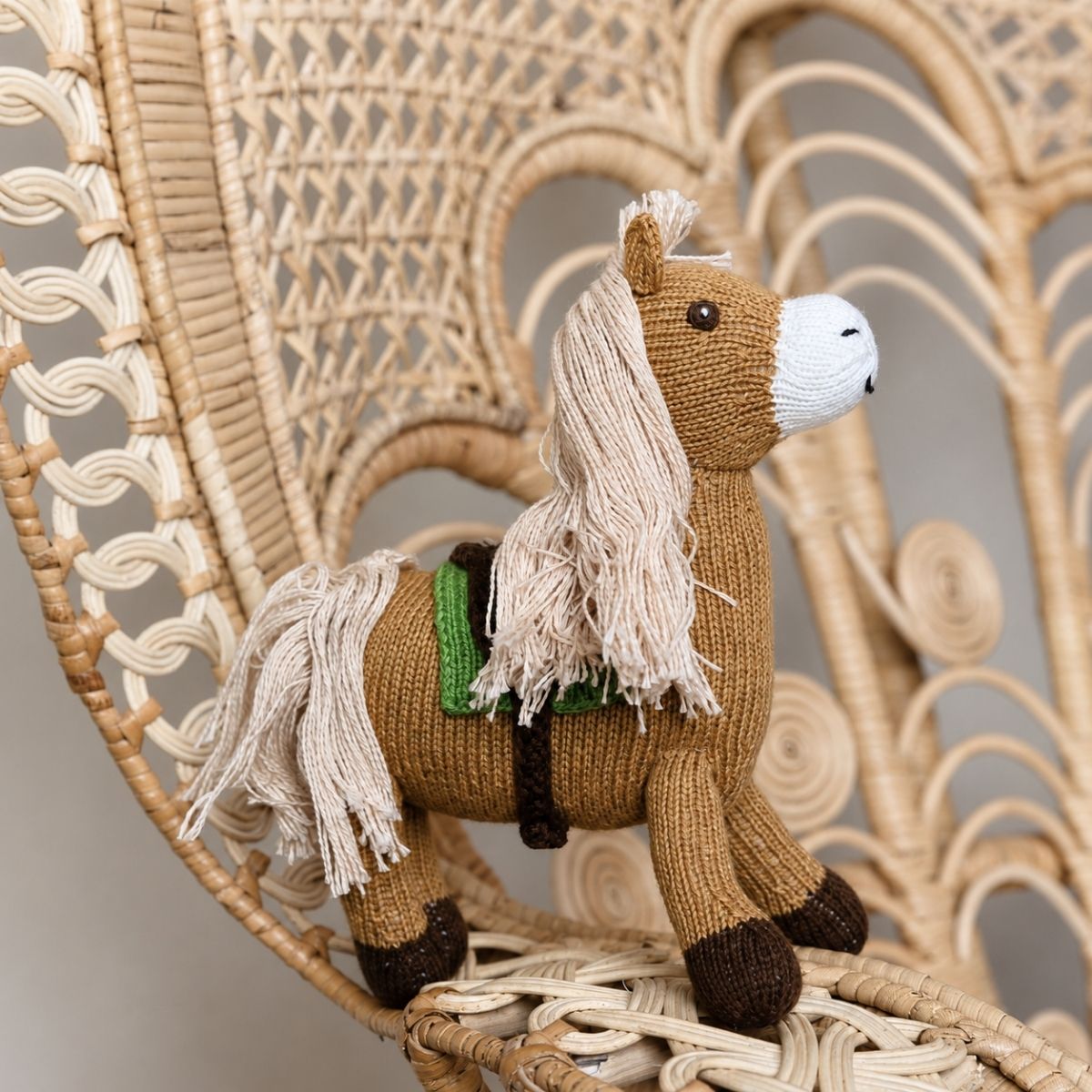 Braunes Bio Kuscheltier Pferd mit heller Mähne und grünem Sattel auf einem Korbstuhl