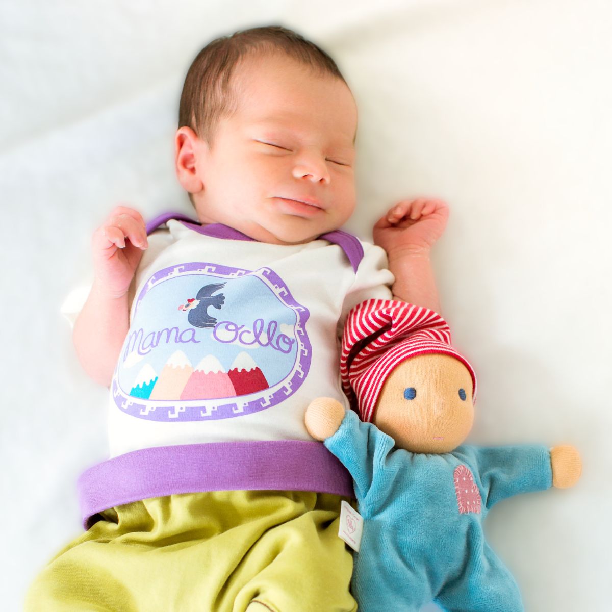 Neugeborenes Baby mit Bio Kurzarmbody in Naturweiß und Lila, liegt auf weicher Decke mit Stoffpuppe