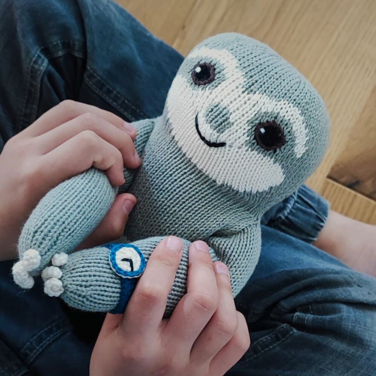 Kind hält ein handgestricktes Bio Faultier Kuscheltier mit blauer Armbanduhr in den Händen