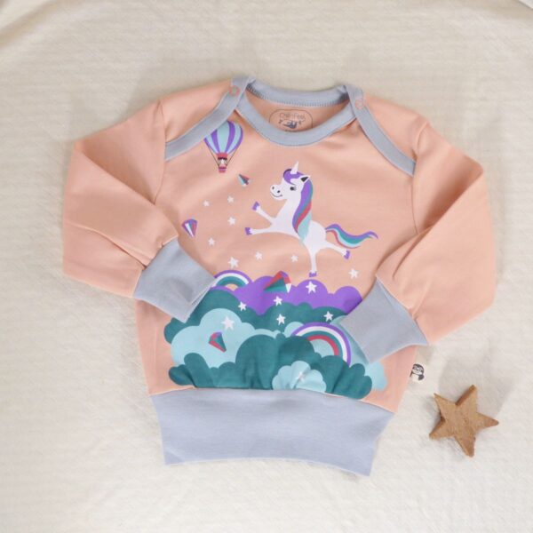 Bio Einhorn Pullover für Babys in Pfirsich mit hellblauen Bündchen, liegt auf beiger Decke mit Deko-Stern