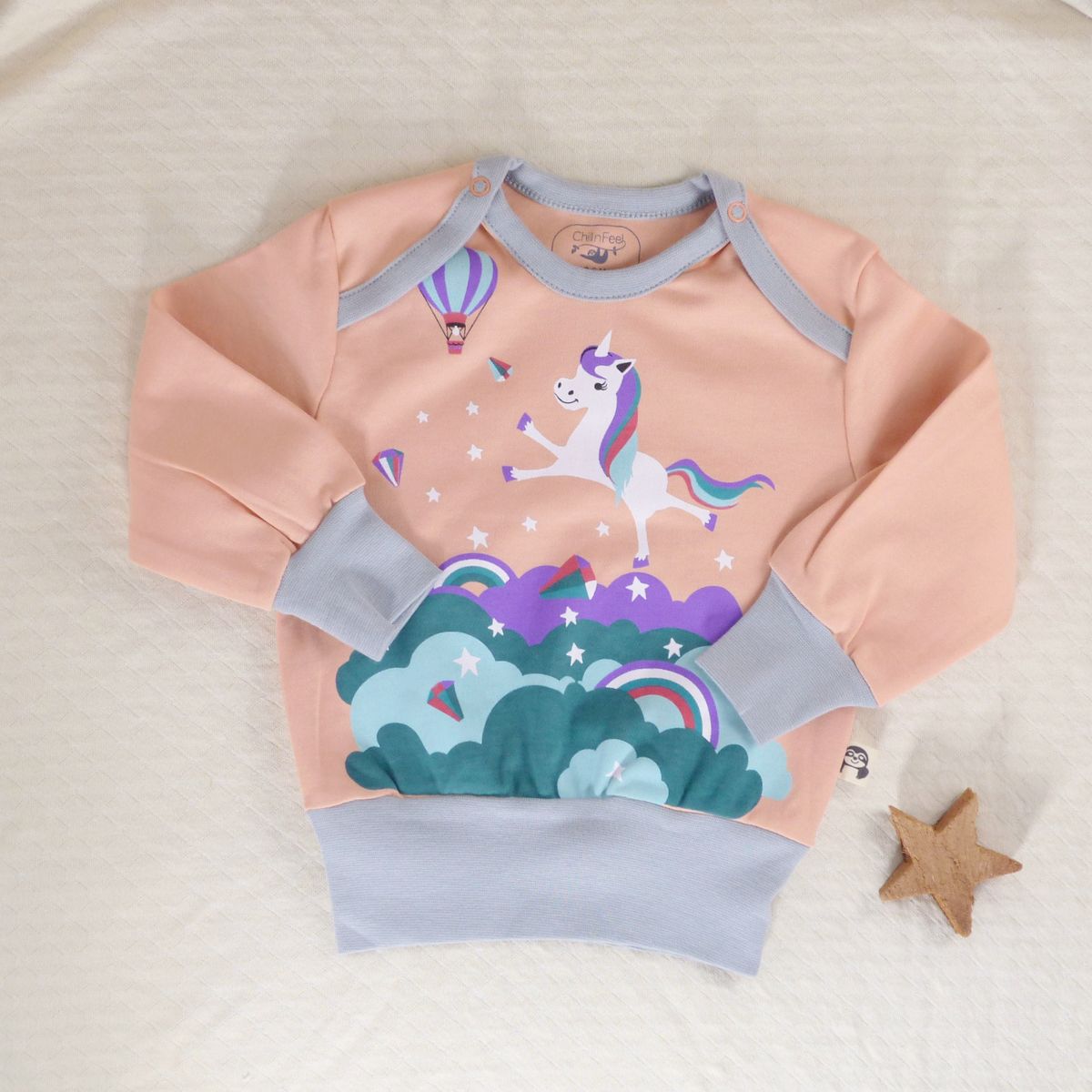 Bio Einhorn Pullover für Babys in Pfirsich mit hellblauen Bündchen, liegt auf beiger Decke mit Deko-Stern
