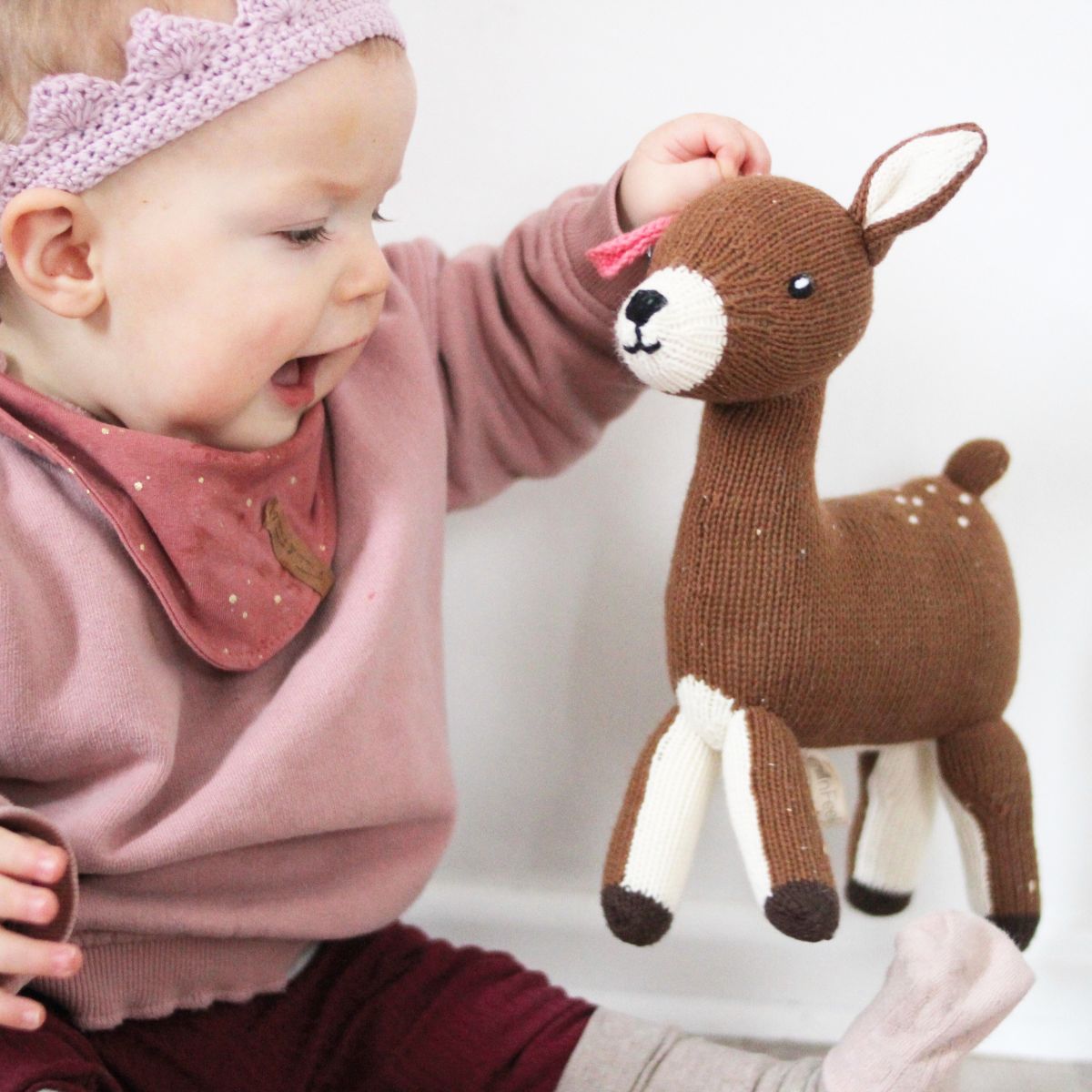 Lächelndes Baby hält ein handgestricktes Bio Kuscheltier Reh aus Baumwolle in der Hand