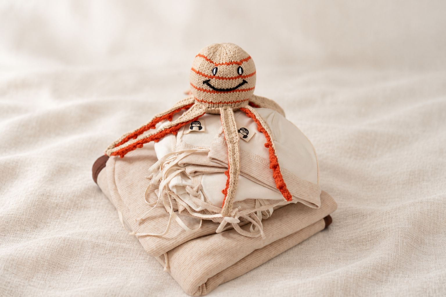 Geschenkidee zur Baby Shower Party: gefaltete Babykleidung in Naturtönen mit einem beige und orange gestrickten Oktopus auf hellem Leinen.