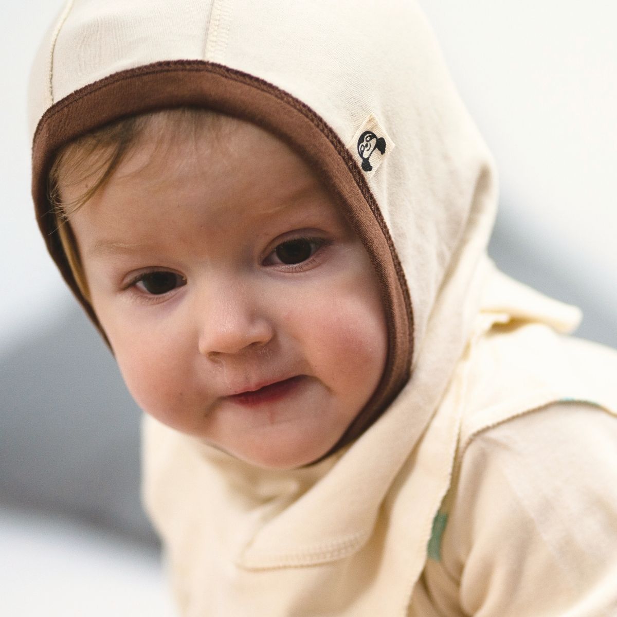 Baby mit beiger Schlupfmütze und braunem Rand aus Bio Pima Baumwolle, hautfreundlich und nahtlos, ideal bei Neurodermitis.