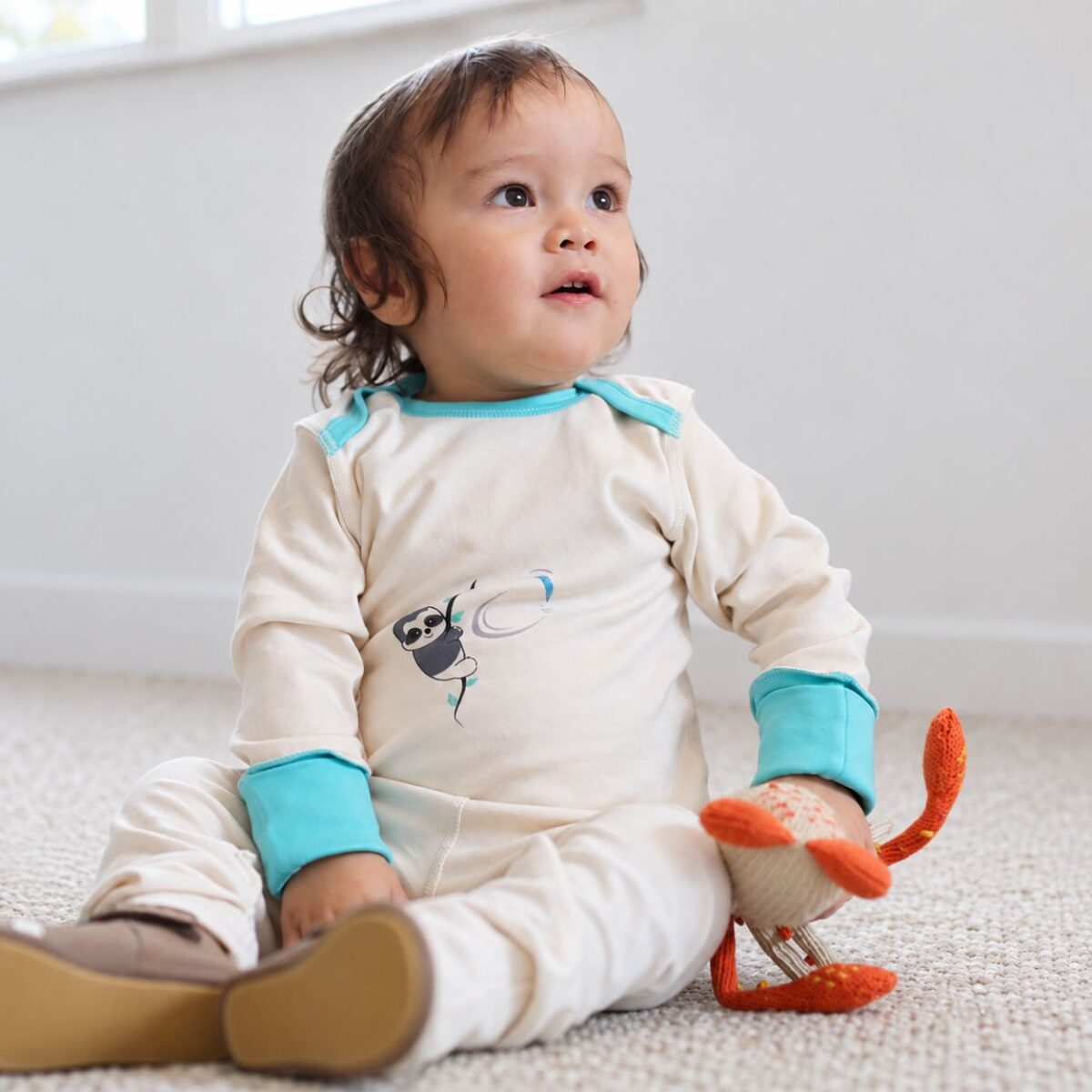 Sitzendes Kleinkind trägt ein cremefarbenes Baby-Langarmshirt mit Kratzschutz, türkisen Bündchen und Faultiermotiv.