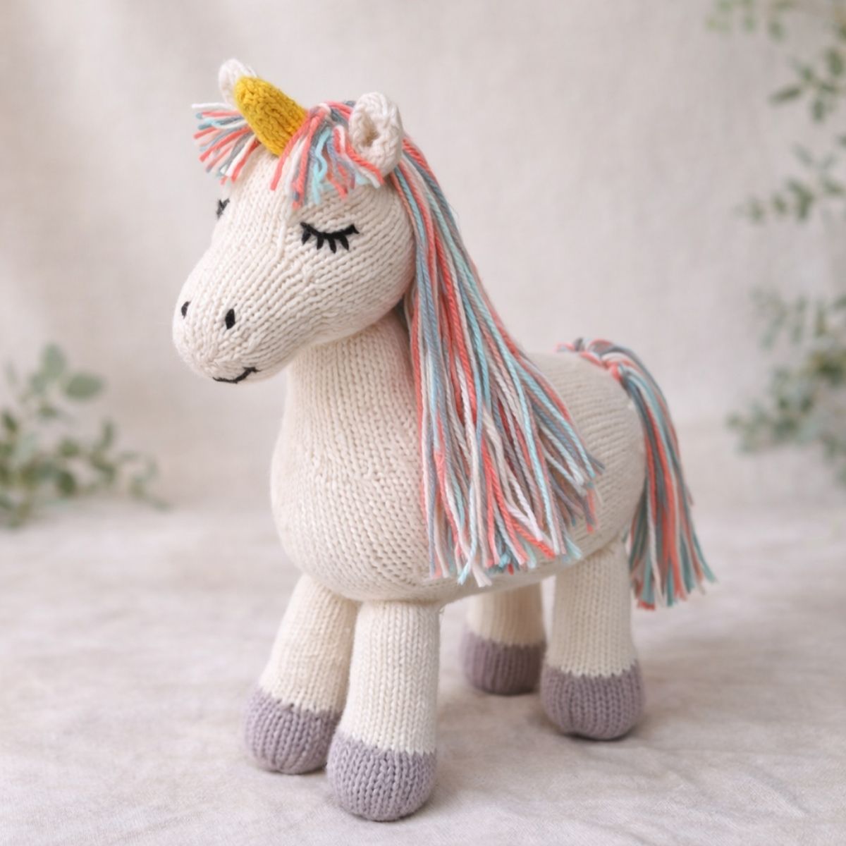 Handgestricktes Kuscheltier Einhorn aus Bio-Baumwolle mit pastellfarbener Mähne auf naturfarbenem Hintergrund