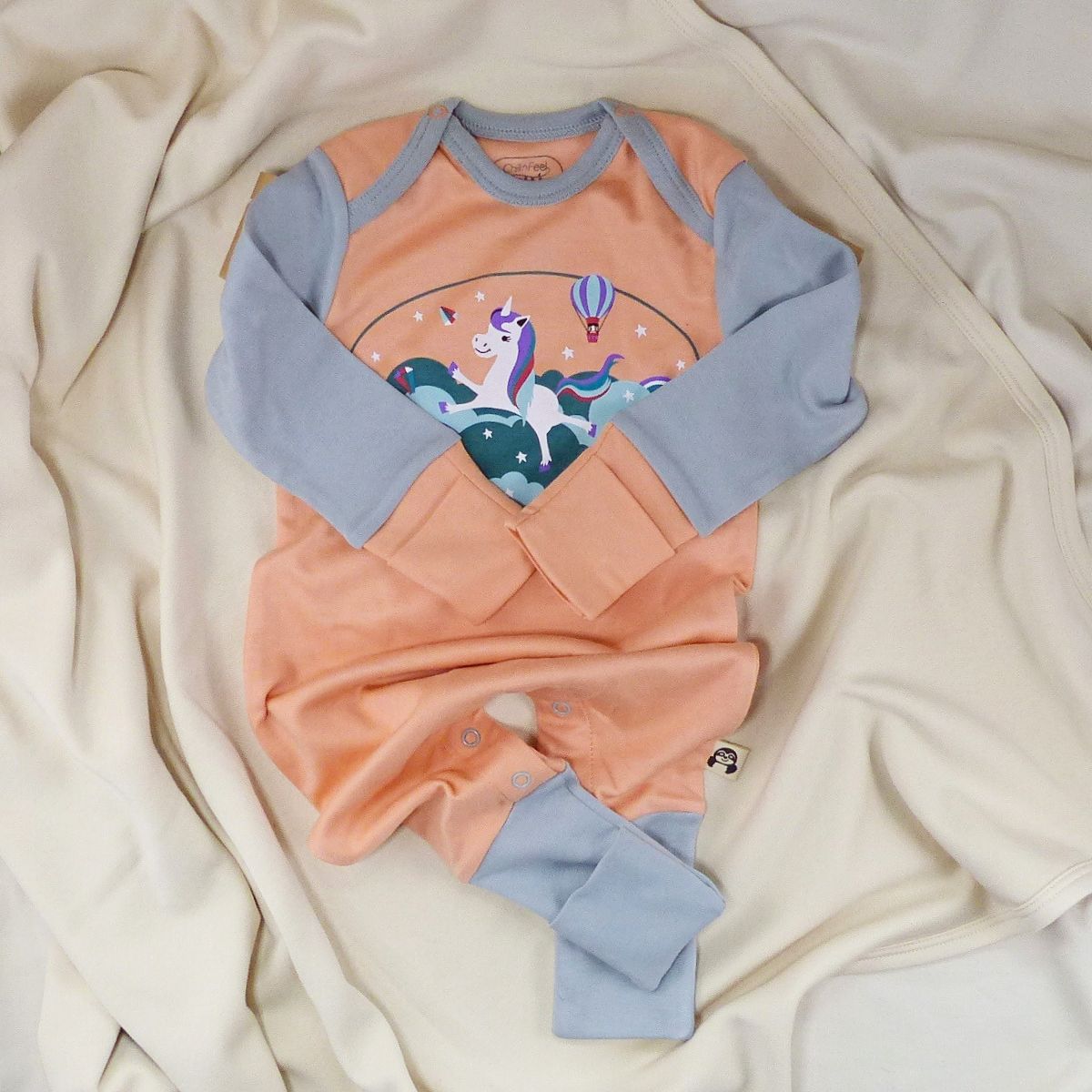 Babystrampler mit Einhorn und graublauen Bündchen, daneben gestricktes Faultier auf heller Decke