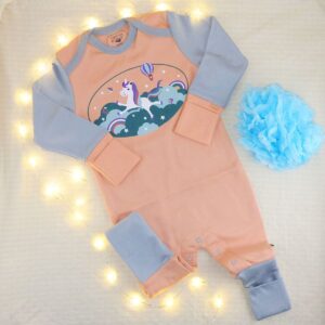 Babystrampler mit Umschlagbündchen in Peach mit Einhorn-Motiv, daneben eine blaue Deko-Blume, eingerahmt von einer Lichterkette