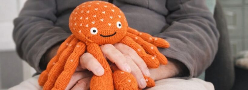 Oranger Kuscheltier-Krebs in den Händen eines älteren Menschen zur sensorischen Stimulation und Unterstützung der Kontrakturprophylaxe bei Demenz