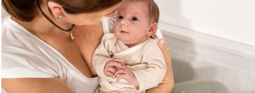 Baby Erstausstattung mit weicher PimaCare Kleidung aus Pima Baumwolle – Neugeborenes geborgen auf dem Arm der Mama.