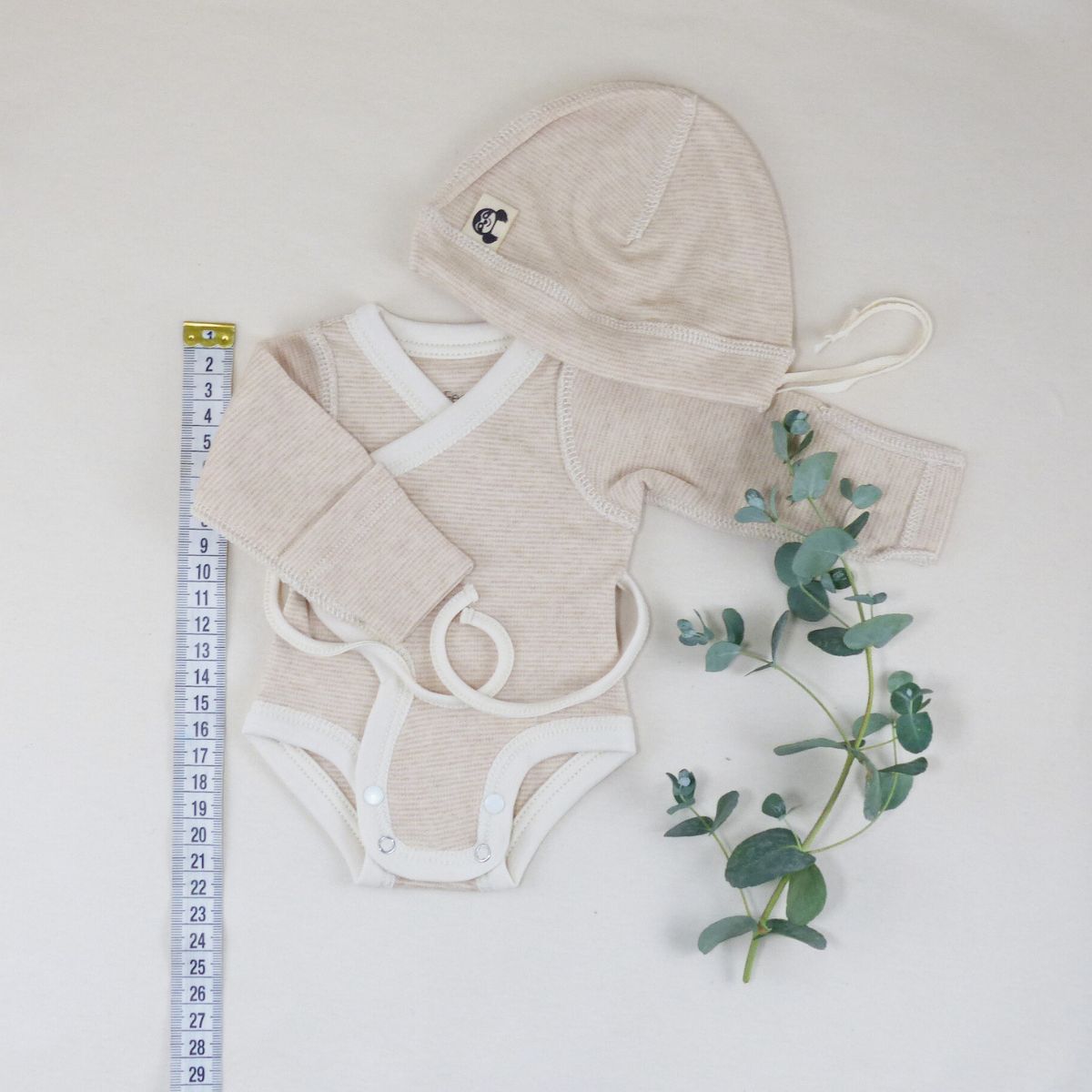 Frühchen Wickelbody in Größe 42, geringelt in Beige-Naturtönen, mit Mütze und Maßband als Größenvergleich, liebevoll arrangiert mit Eukalyptuszweig auf hellem Hintergrund