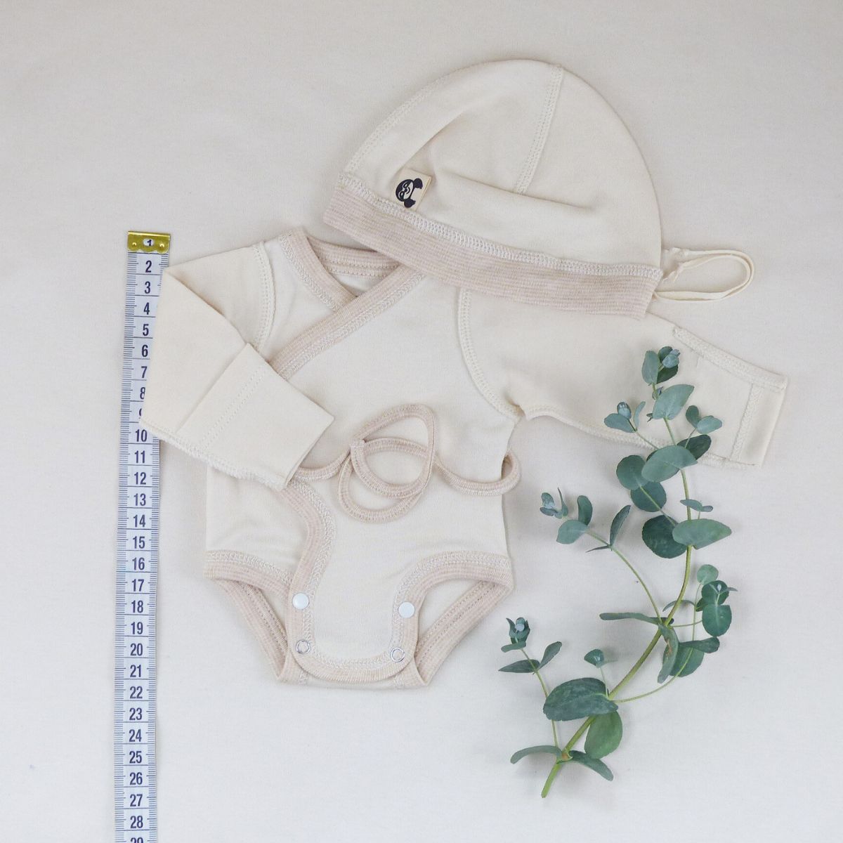 Frühchenkleidung Größe 42 – naturbeiges Set aus Wickelbody und Mütze aus GOTS-zertifizierter Bio-Baumwolle, daneben ein Maßband und Eukalyptuszweig zur sanften Dekoration