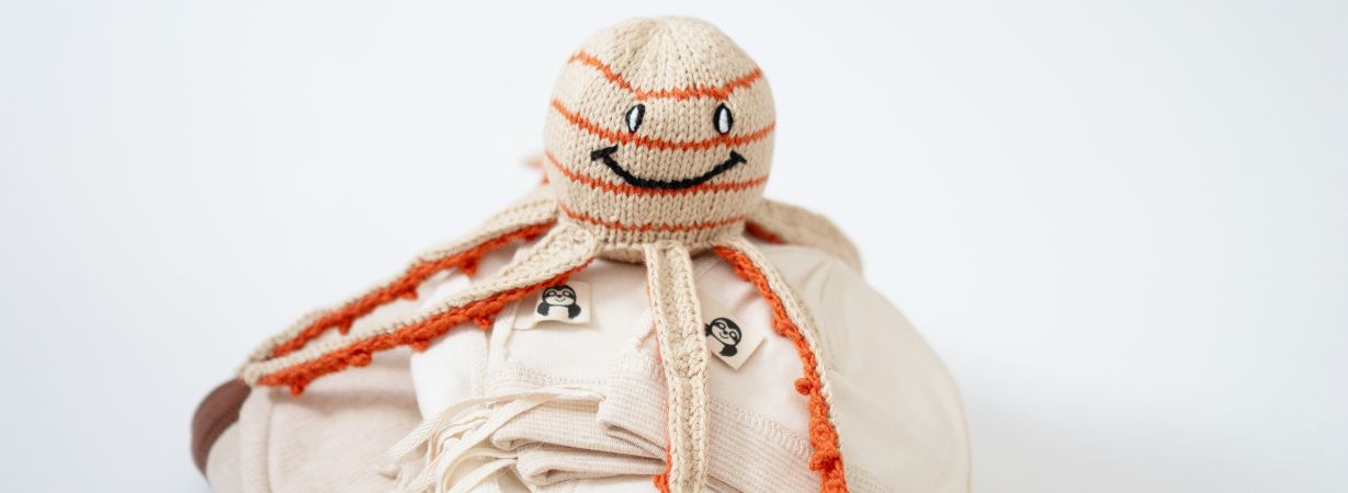 Ein gestrickter Oktopus liegt auf sorgfältig gefalteter Frühchenkleidung aus Bio-Baumwolle – Symbol für Geborgenheit und die liebevolle Erstausstattung, die über die Krankenkasse erhältlich ist.