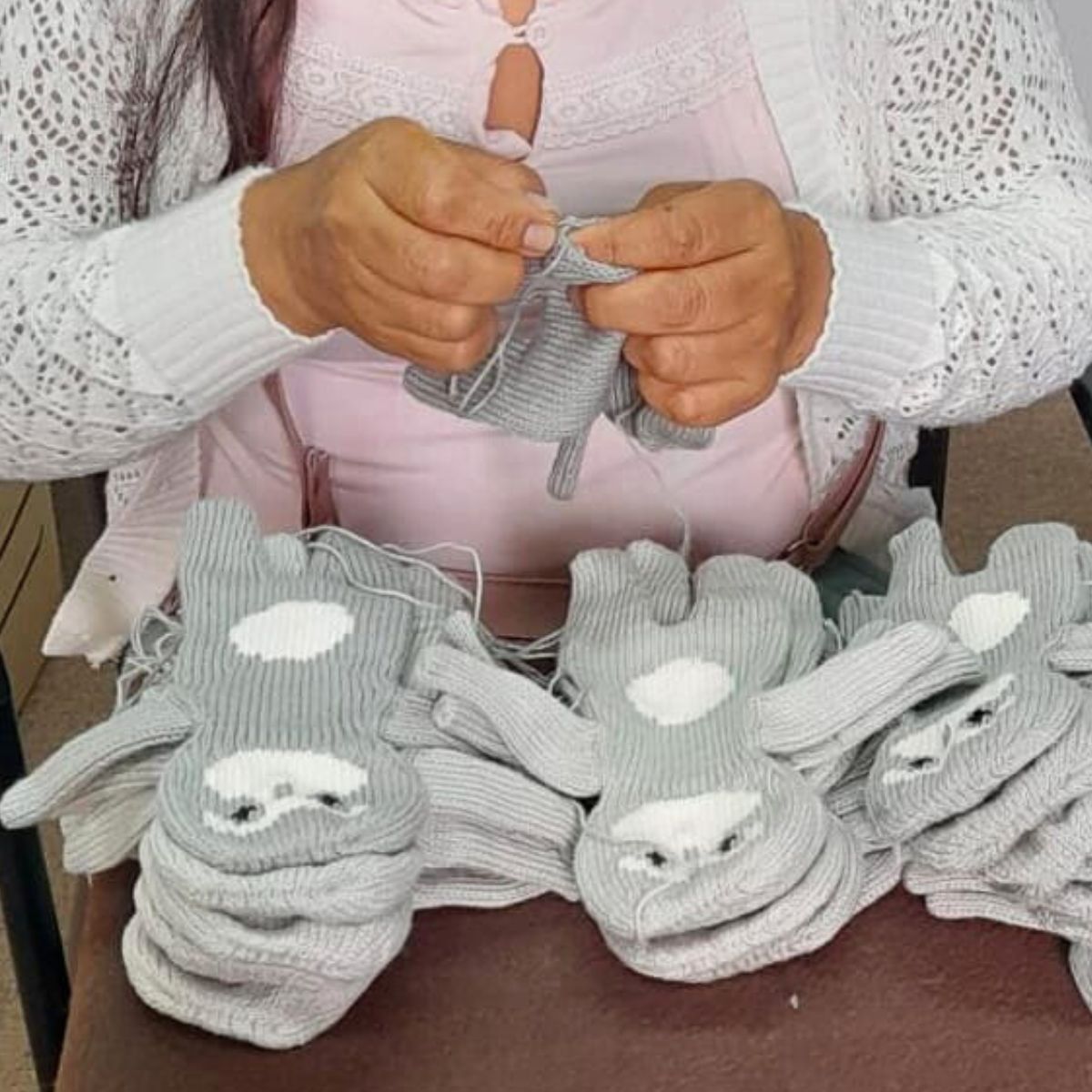 Frau strickt in liebevoller Handarbeit graue gestrickte Kuscheltiere in Faultierform; mehrere ungestopfte Kuscheltierkörper liegen gestapelt vor ihr.