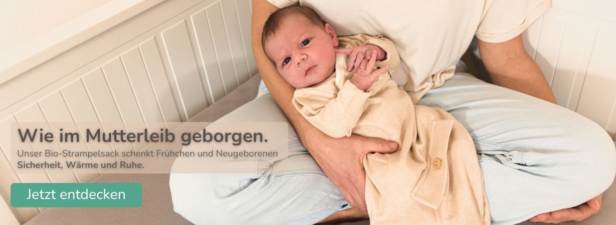 Ein Neugeborenes liegt geborgen im Arm eines Elternteils und trägt einen Strampelsack aus GOTS-zertifizierter Bio-Pima-Baumwolle. Der Strampelsack schenkt Frühchen und Neugeborenen Wärme, Sicherheit und Geborgenheit – wie im Mutterleib.