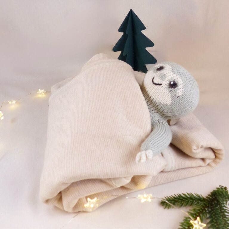 Bio Babydecke aus Pima Baumwolle als nachhaltiges Weihnachtsgeschenk, liebevoll inszeniert mit Lichterkette, kleinem Tannenbaum und Faultier-Kuscheltier.