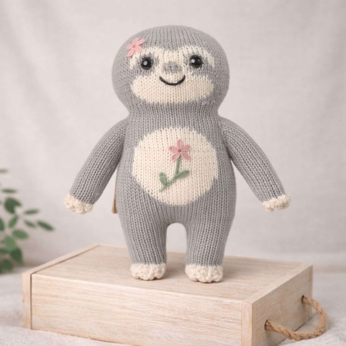 Handgestricktes Faultier Stofftier Blumen-Fauli mit rosa Blume in sanfter, minimalistischer Umgebung