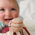 Fröhliches Neugeborenes im Sommer trägt leichte Bio-Pima-Baumwollkleidung und liegt mit einem gestrickten Oktopus auf einem weißen Bett.