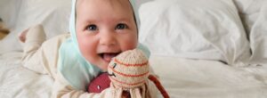 Fröhliches Neugeborenes im Sommer trägt leichte Bio-Pima-Baumwollkleidung und liegt mit einem gestrickten Oktopus auf einem weißen Bett.