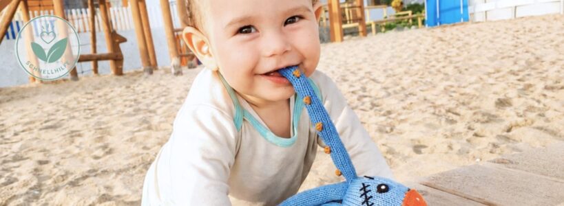 Fröhliches Baby spielt am Strand im Urlaub mit Neurodermitis bei Sonne, Sand und Meer