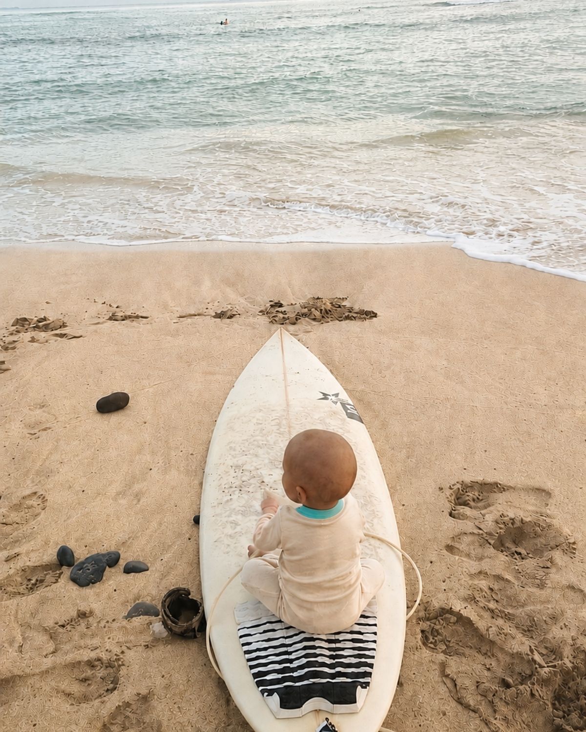 Baby in naturbeiger Kleidung mit blauer Einfassung sitzt am Strand auf einem Surfbrett und blickt auf das Meer.
