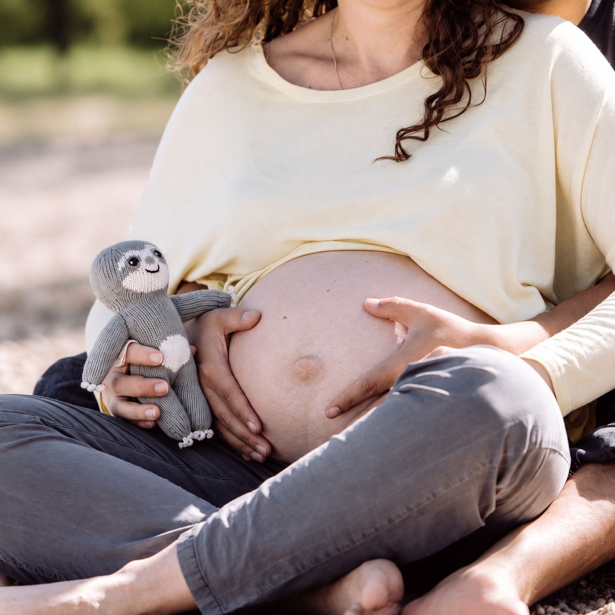 Schwangere hält das gestrickte Baby-Faultier Chilly neben ihrem Babybauch, die Hände ihres Partners liegen am Bauch.